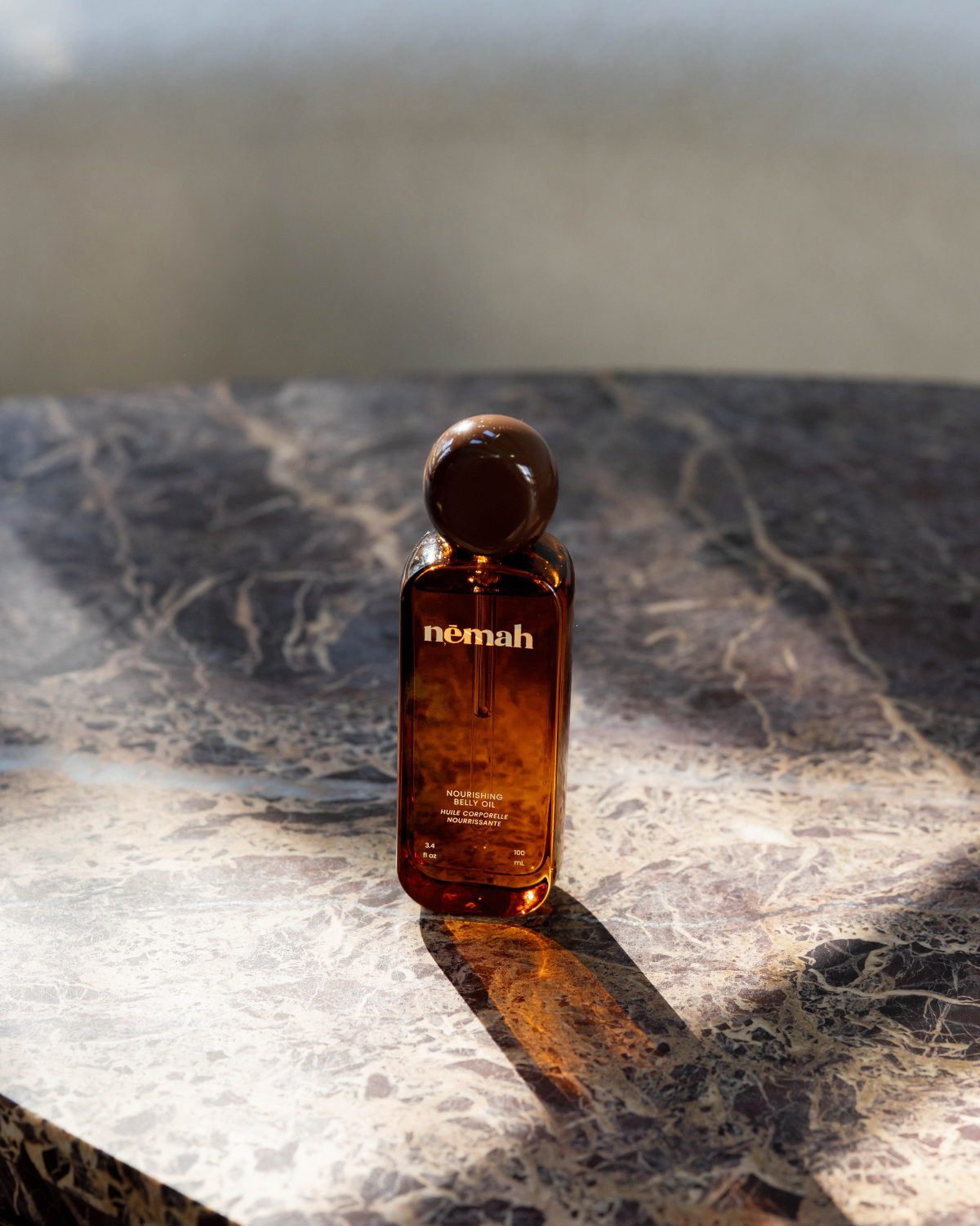A brown glass bottle of Nourishing Belly Oil sits on a sunlit dark marble, casting a shadow. The modern design and amber hue highlight its plant-based squalane, emphasizing the nourishing qualities of this product.