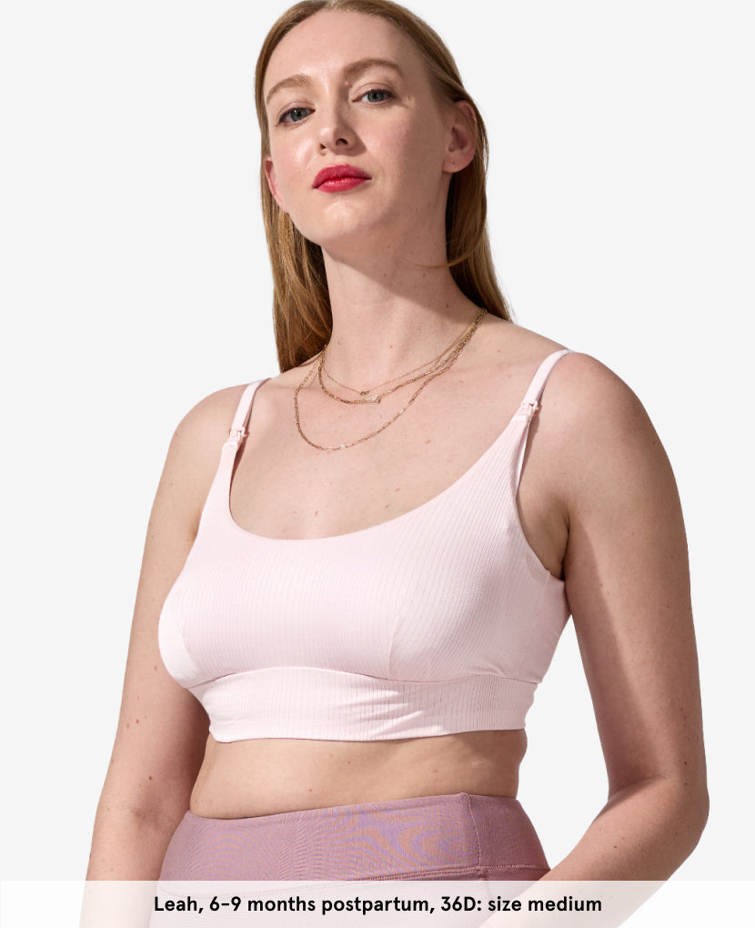A woman with fair skin and long red hair wears The Everything Bra in light pink and mauve leggings, standing against a plain background. Text below reads: Leah, 6–9 months postpartum, 36D; size medium.