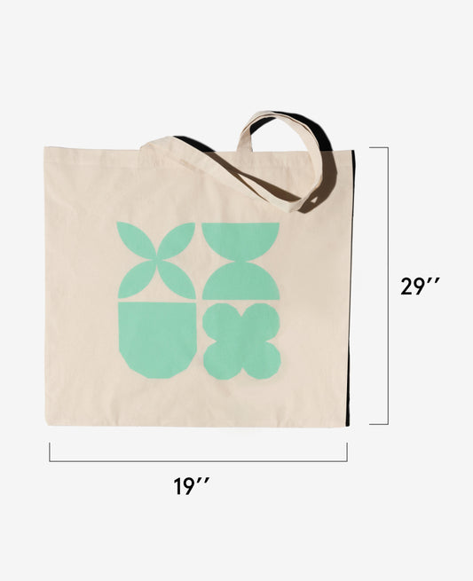 The Hospital-to-Playground Tote is a beige washable bag featuring green geometric shapes, perfect for hospital stays or postpartum recovery. It measures 19" wide by 29" tall and has two handles—one flat, one bent.