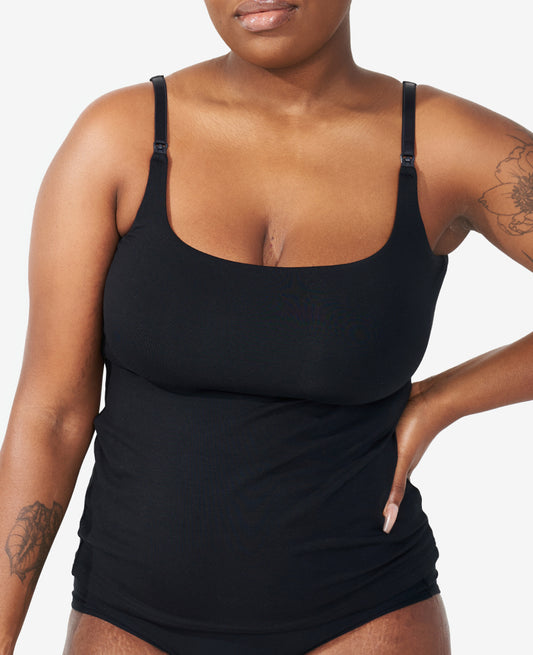 A woman wearing a black tank top with thin straps and matching underwear stands with one hand on her hip. She has floral tattoos on her shoulder and forearm. The background is plain white.