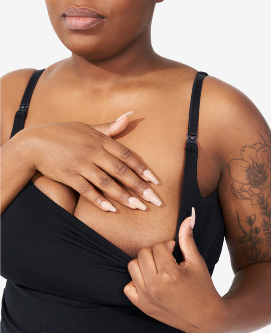 A person with long, manicured nails unclips the strap of a black nursing tank top, pulling down the fabric to reveal part of their chest; a floral tattoo is visible on their upper arm.