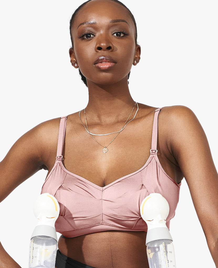 A woman uses The All-Needs Bundle • 20% off, featuring a pink nursing bra and dual electric breast pumps with bottles, highlighting essential postpartum support on a plain white background.
