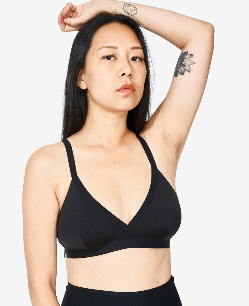 A woman with long black hair and visible tattoos poses with one arm raised, wearing The All-Needs Bundle • 20% off: a black nursing bra and high-waisted black bottoms, against a plain white background.