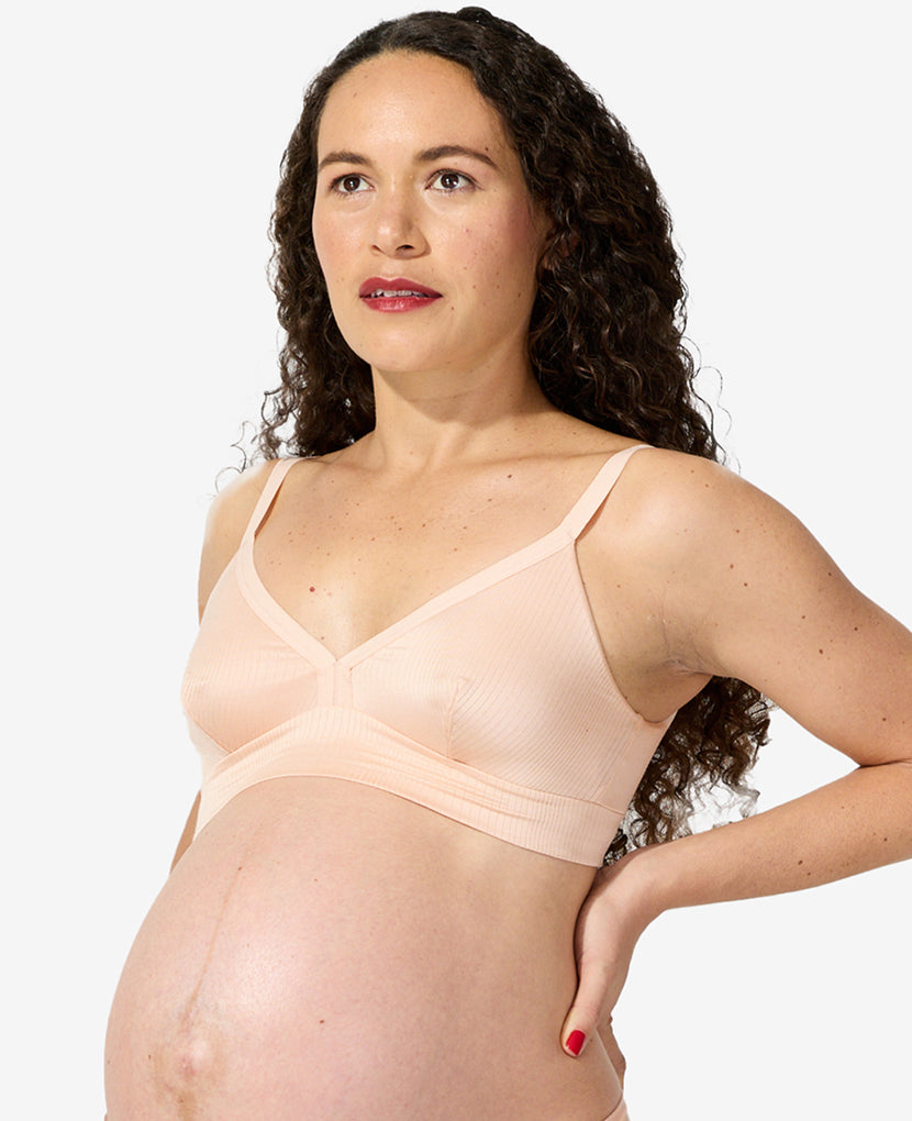 A pregnant woman with long curly hair wears The All-Needs Bundle (20% off), featuring a light peach nursing bra, while standing against a plain white background and gazing slightly upward and to the side.