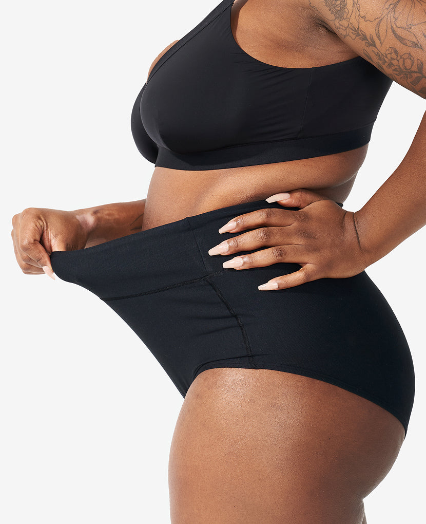 A person stretches the waistband of The All-Needs Bundle • 20% off—featuring a black nursing bra and high-waisted black underwear—demonstrating their flexibility and comfort against a white background.