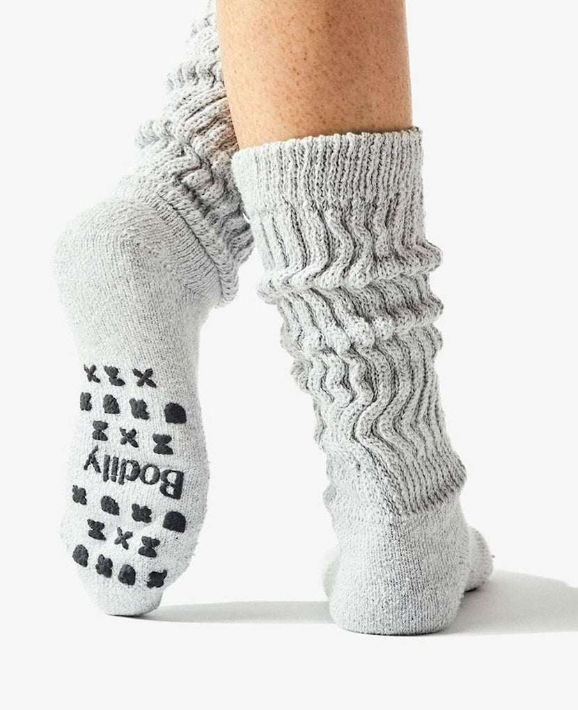 Someone stands on one foot with the other heel raised against a plain white background, wearing Cozy Socks: Grey—light gray, breathable cotton grip socks featuring no-slip sole dots and the “Bodily” logo.