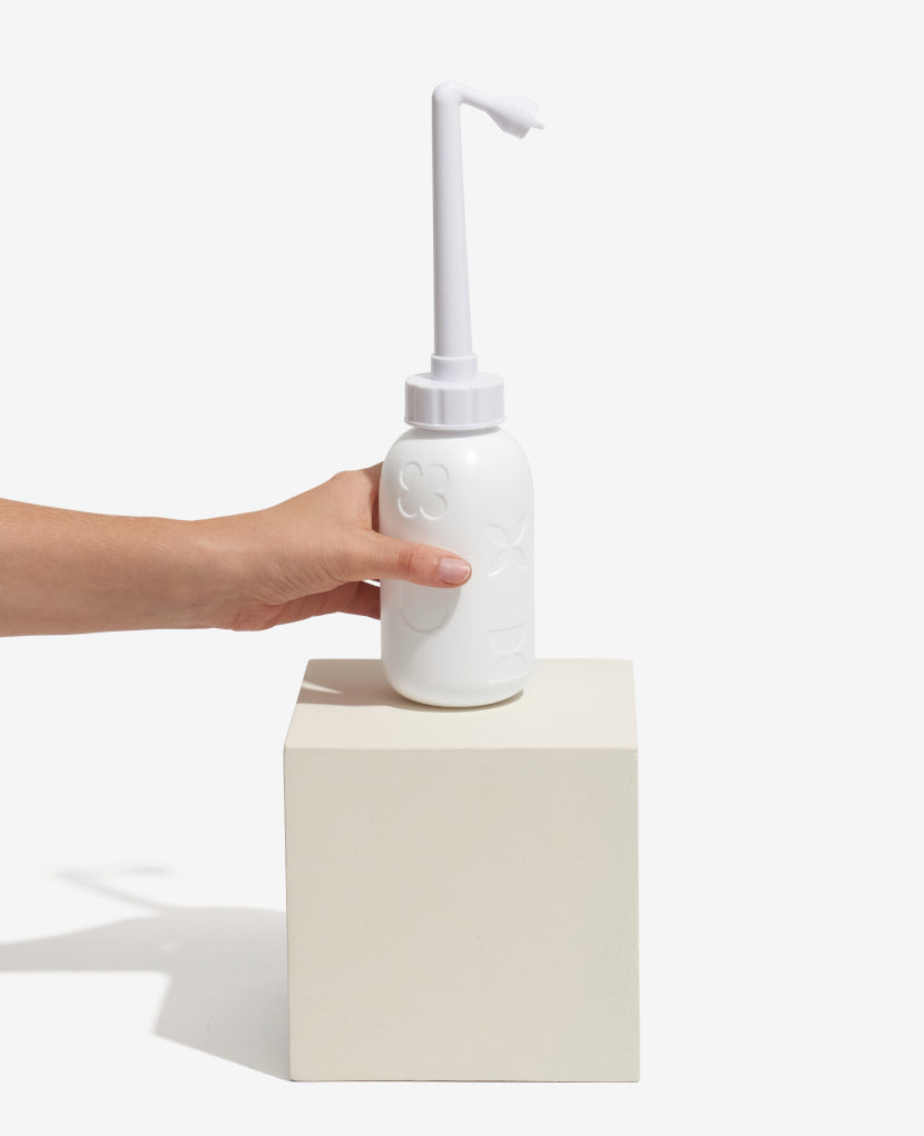 A hand holds the Peri Wash Bottle, a white postpartum perineal care bottle with a long angled spout, placed on a white rectangular block against a plain white background.