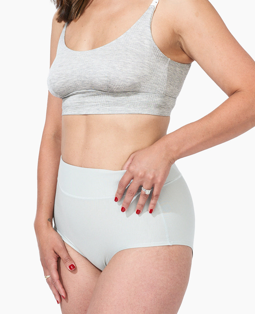 Woman wears The All-In Panty: 3-Pack • Save 16%—a grey high-waisted panty and sports bra set. She poses confidently against a white background, showcasing these comfy postpartum underwear essentials.