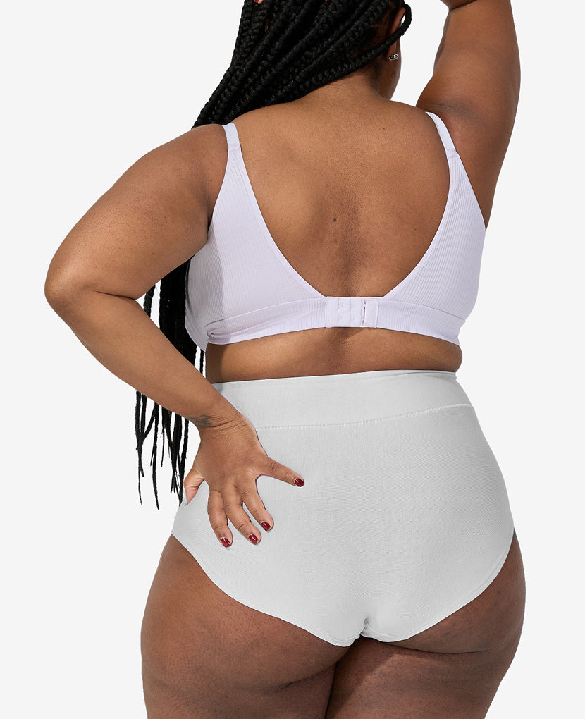 A person with long braids is seen from behind, wearing The All-In Panty: 3-Pack • Save 16% in white, paired with a matching bra, standing against a plain light background with one arm raised and the other on their hip.