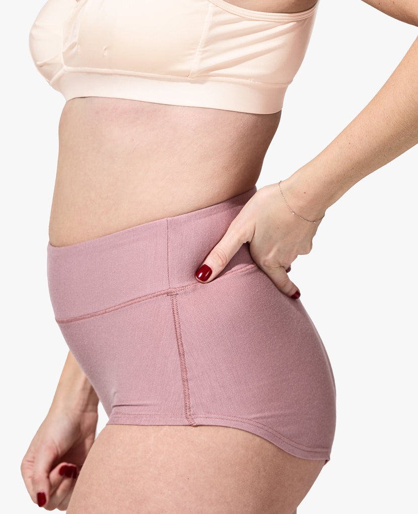 A person stands in profile against a white background wearing The All-In Panty: 3-Pack • Save 16%, featuring a light pink bra and mauve high-waisted panty, with one hand on their hip.