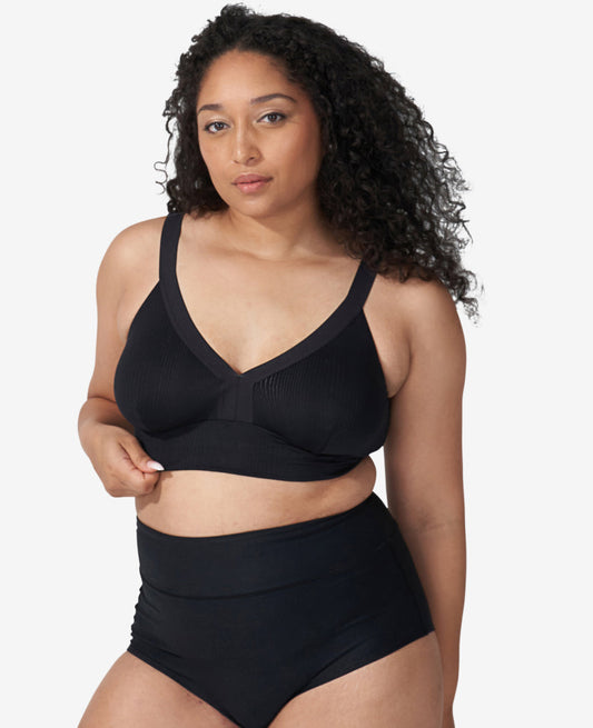 A woman with long curly hair stands confidently against a plain light background, wearing The Effortless Bra: 3-Pack • Save 20%, showcasing stylish and comfortable black nursing bras for postpartum wear.