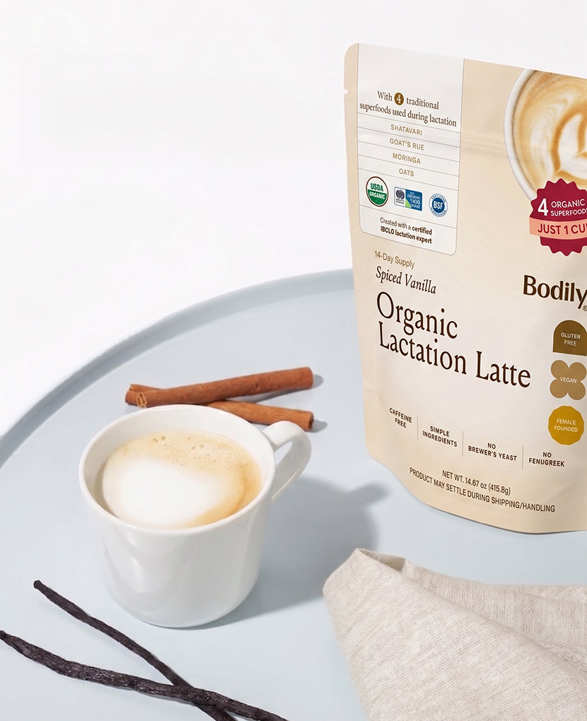 A cup of frothy latte sits beside cinnamon sticks, vanilla beans, a beige napkin, and a package of Organic Lactation Latte: Single in spiced vanilla—ideal gentle lactation support for breastfeeding moms.