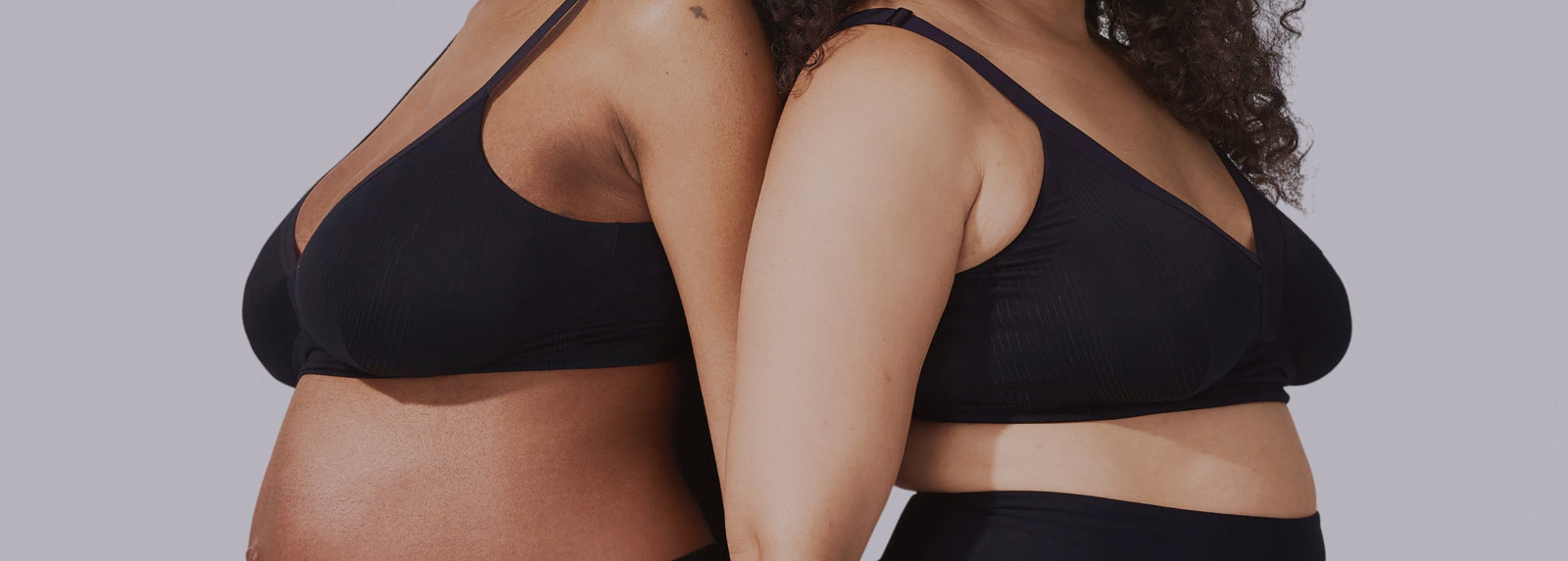 Two women wearing black bras and underwear stand back-to-back, showing their midsections. Both have visible curves and stretch marks, highlighting body positivity and diversity. The background is a plain, light color.