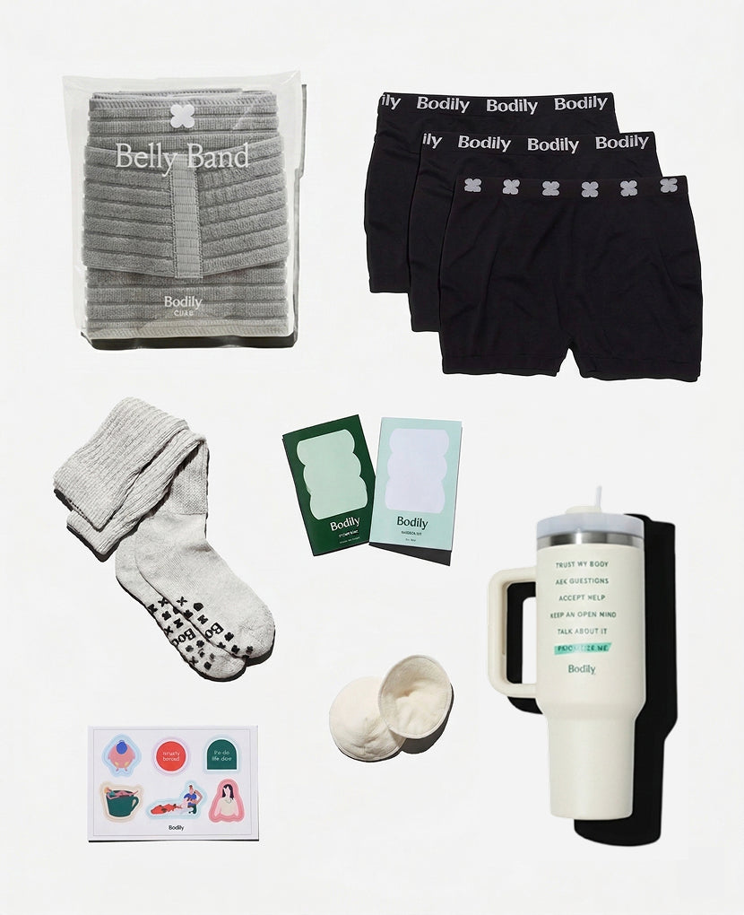 A flat lay shows the Scheduled C-Section Bundle a belly band in packaging, three pairs of black Bodily underwear, grey socks, nipple pads, cards, a travel mug, and colorful stickers—postpartum essentials for recovery.