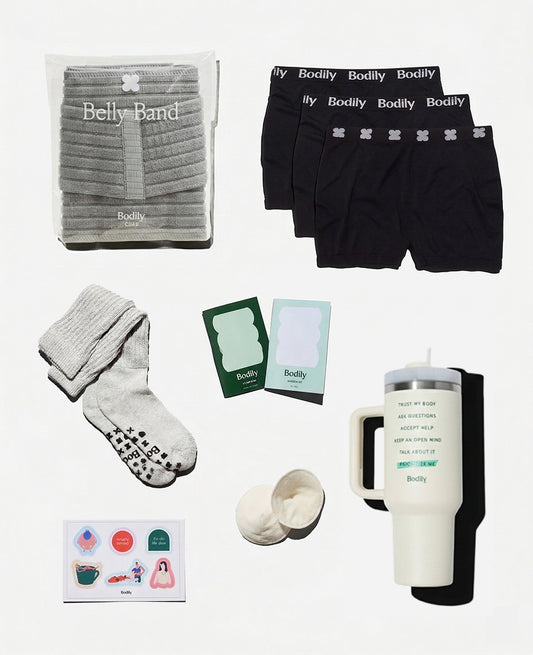 A flat lay shows the Scheduled C-Section Bundle a belly band in packaging, three pairs of black Bodily underwear, grey socks, nipple pads, cards, a travel mug, and colorful stickers—postpartum essentials for recovery.